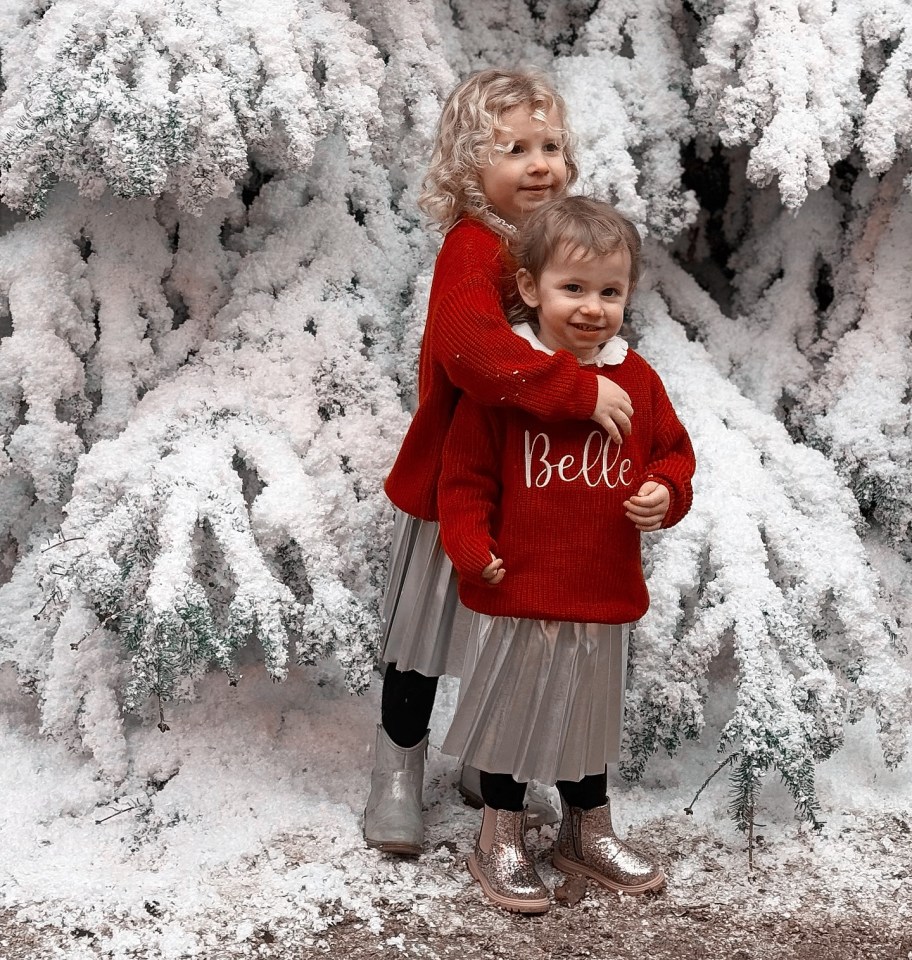 Two girls in red sweaters hug in front of a snow-covered tree.