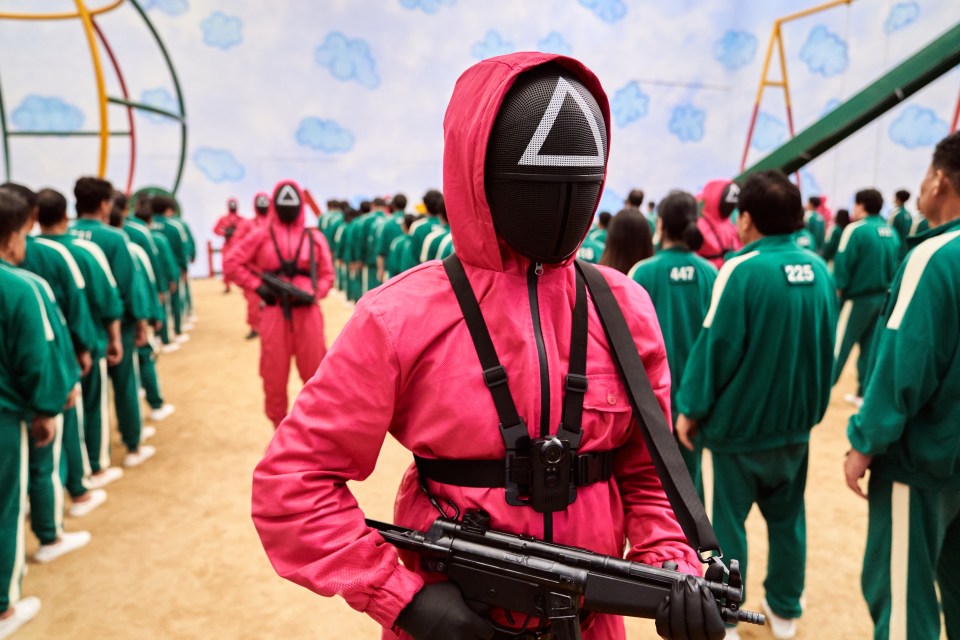 Squid Game guard in pink jumpsuit and triangular mask holding a submachine gun.