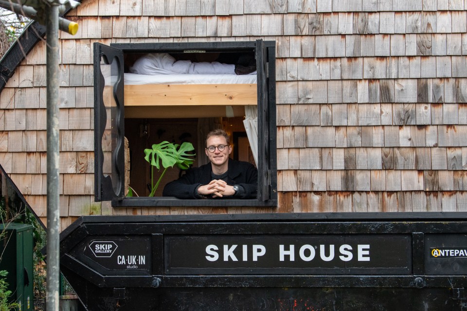 Man smiling in window of small house built from a converted skip.