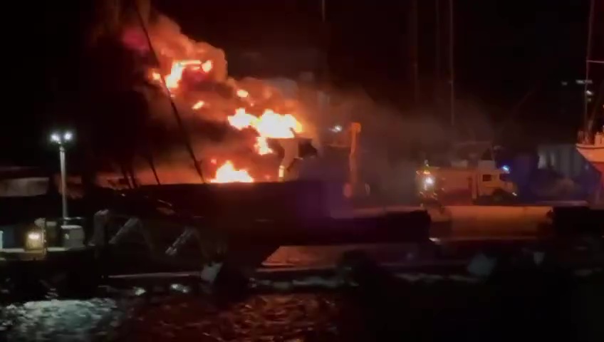 A large superyacht engulfed in flames in a port.