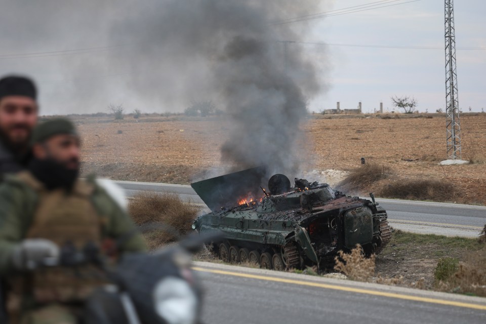 Syrian opposition fighters drive past a burning armored vehicle