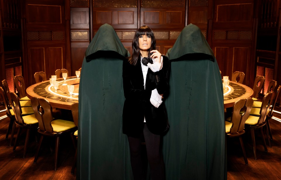 Claudia Winkleman stands between two figures in cloaks at a large round table.