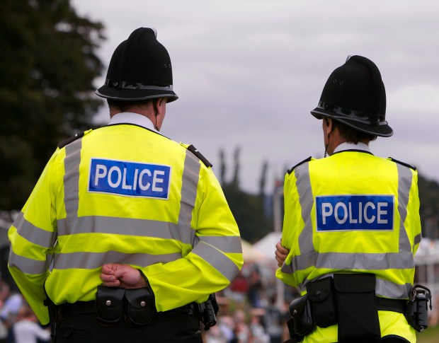 Two British police officers in reflective vests and yellow hard hats guard the entrance to an arena at a summer fair.