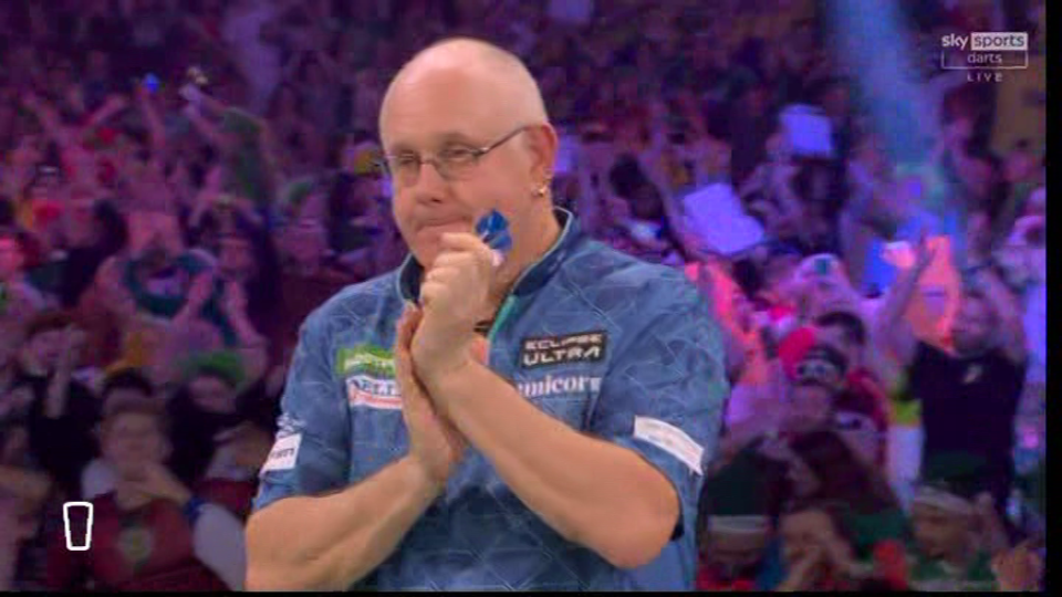 A darts player in a blue shirt applauds in front of a cheering crowd.