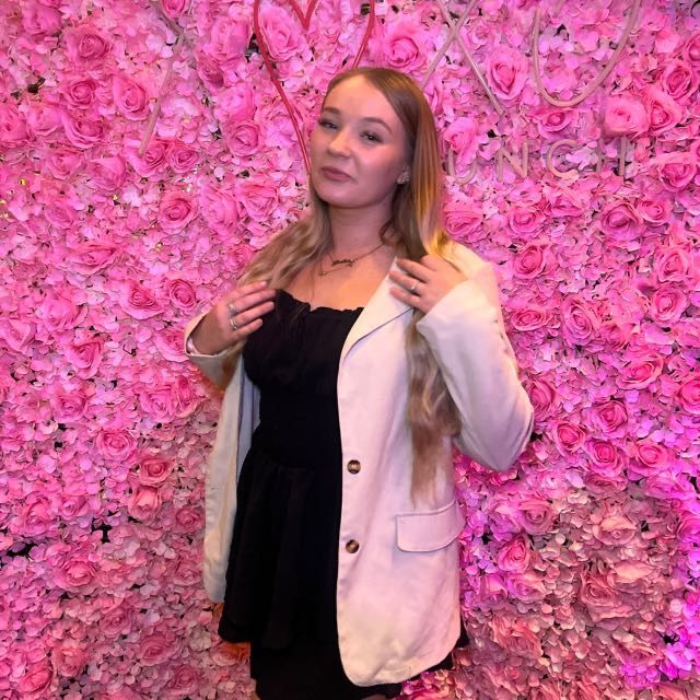 Woman in beige blazer posing in front of pink flower wall.