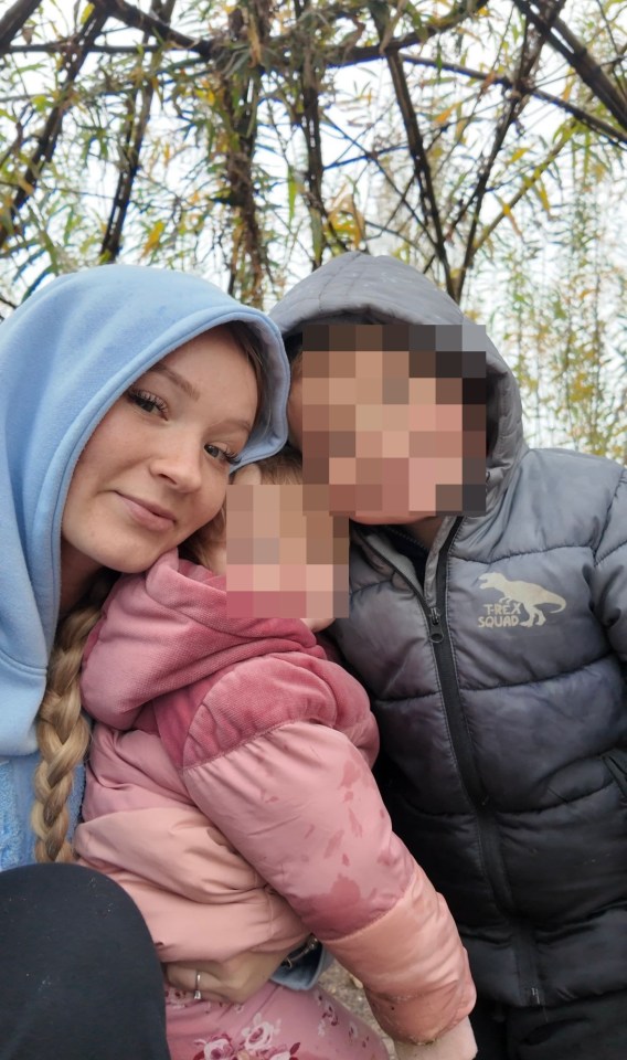 A woman holds a young child while standing next to another child.  The faces of the children are blurred.