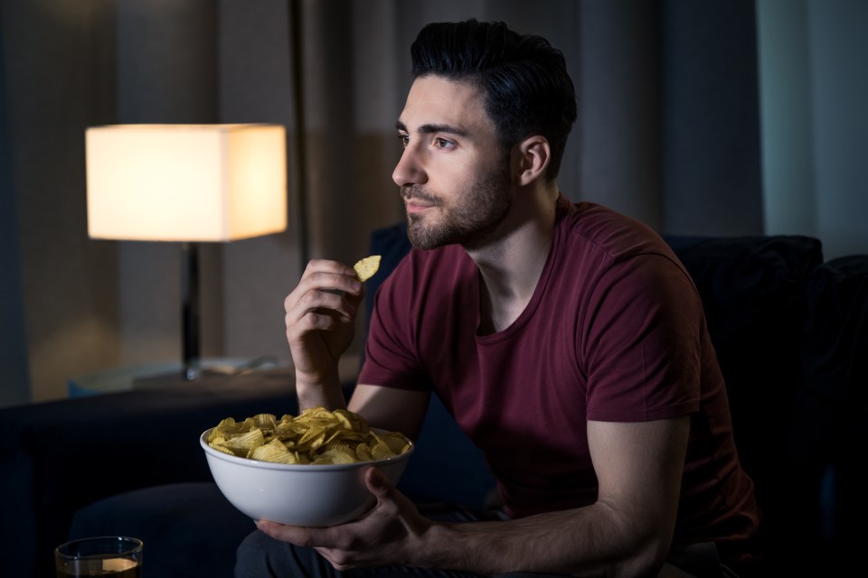 A man relaxing on his sofa, eating chips while watching TV.