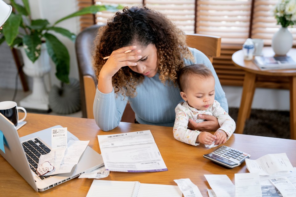 A stressed young mother holding her baby while looking at bills and using a calculator.