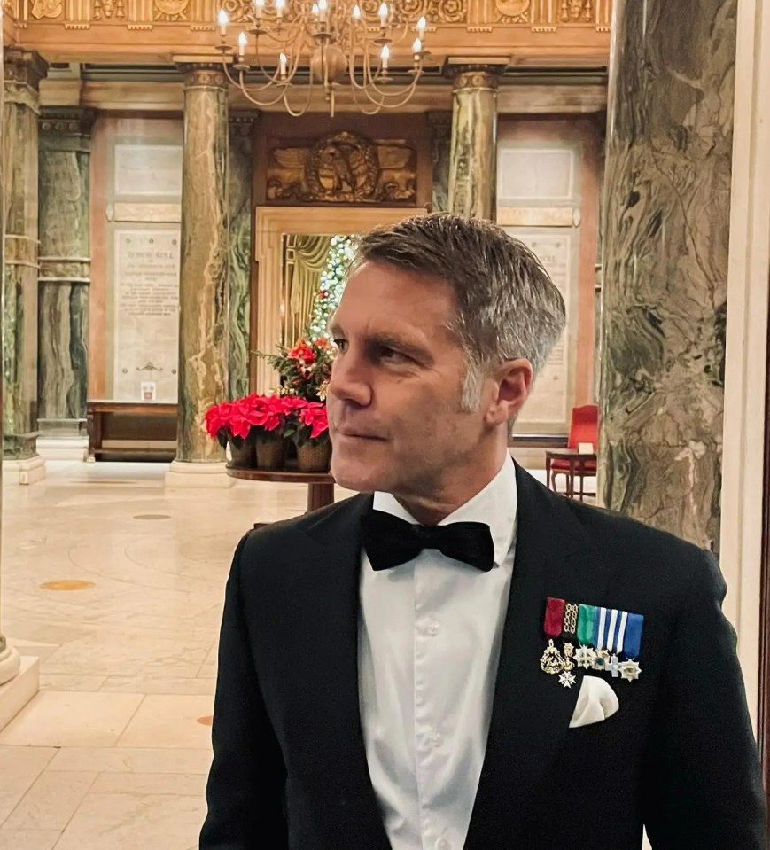 Man in tuxedo and medals in ornate hallway.