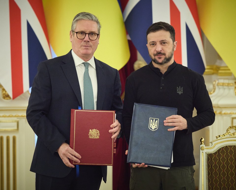 Two men sit at a table, one in a suit, the other in casual clothes; flags of the UK and Ukraine are visible in the background.