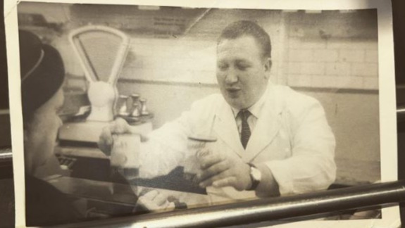 Black and white photo of a man in a white coat behind a counter, appearing to serve a customer.