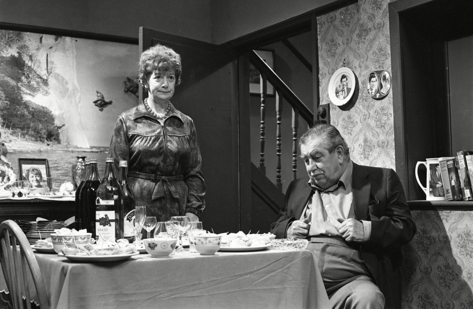 Black and white photo of Hilda and Stan Ogden from Coronation Street, sitting at a table with food and drinks.