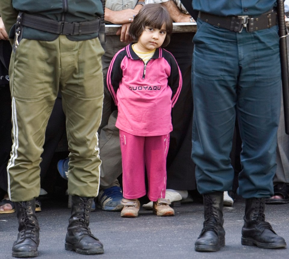 A young girl stands between two uniformed officers at a public execution in Iran.