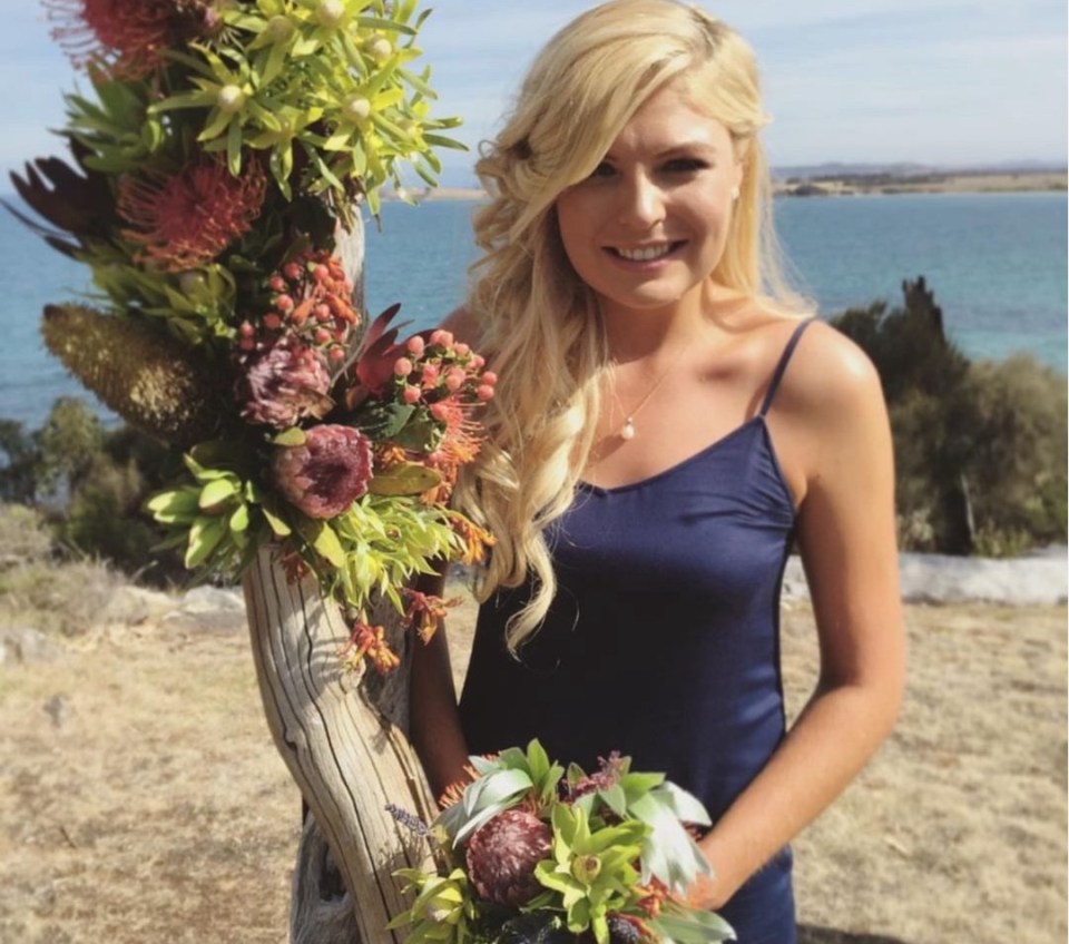 Woman in blue dress holding a bouquet, standing by a floral arrangement.