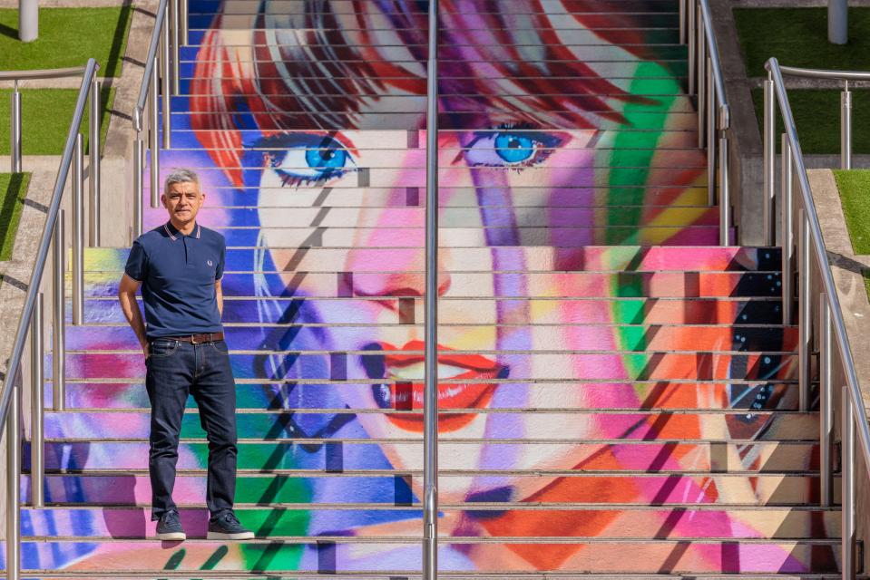 Sadiq Khan standing on stairs with a mural of a woman's face.