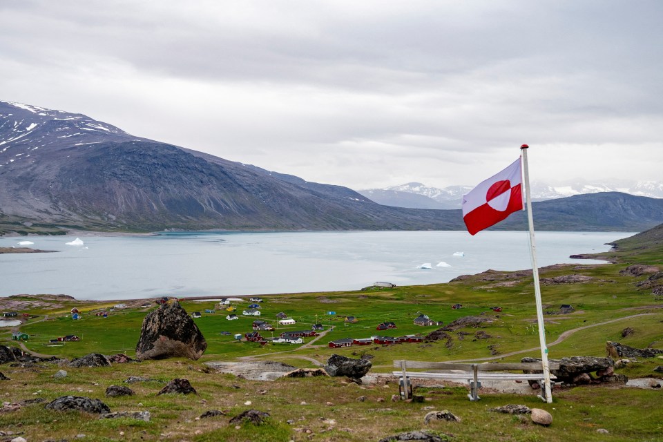 Greenlandic flag flies over a village by the water, with mountains in the background.