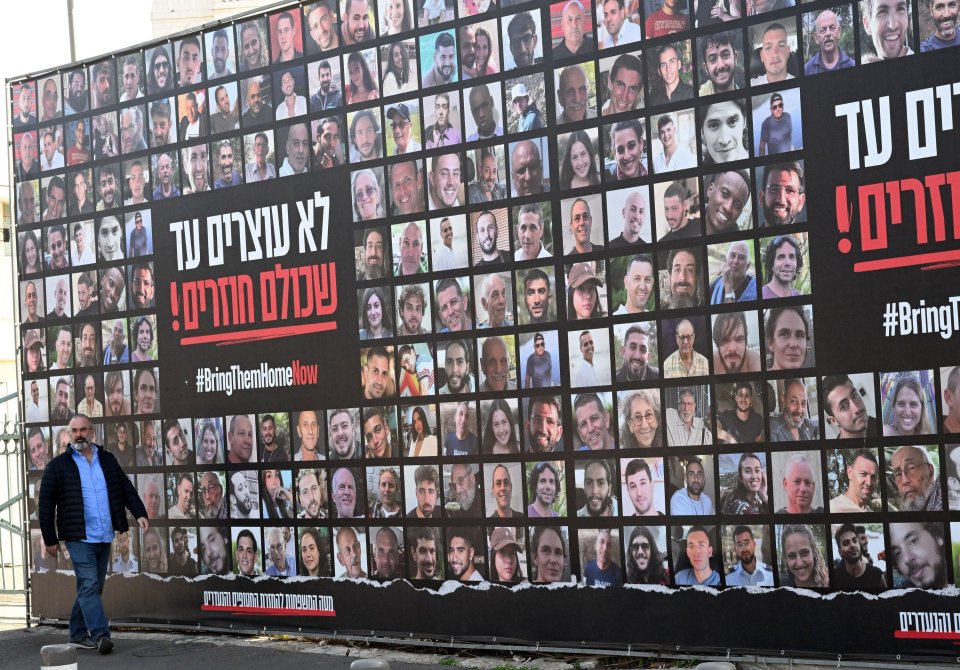 A woman walks past a billboard displaying photos of missing people with the text "Bring them home now" in English and Hebrew.