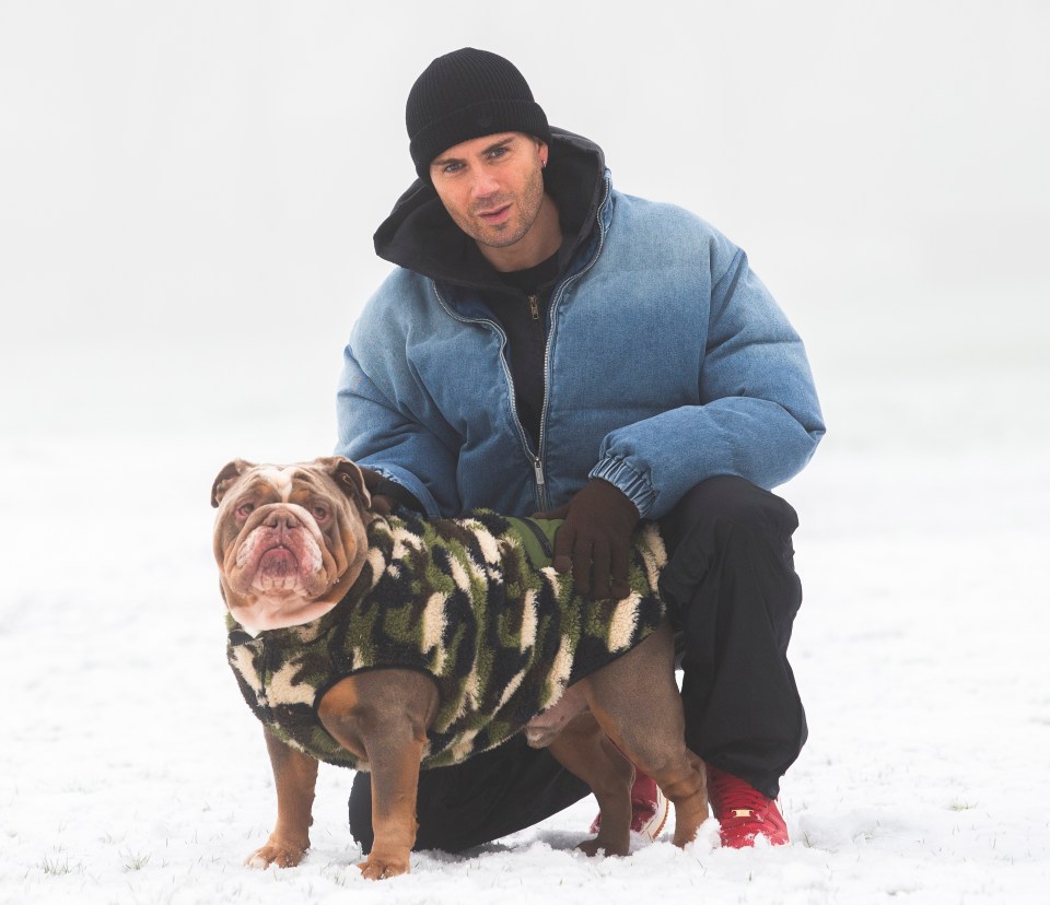 Man kneeling in the snow with his bulldog wearing a camouflage jacket.