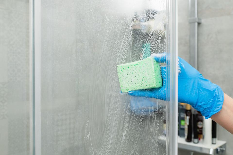 A hand in a blue glove cleaning a shower door with a green sponge.