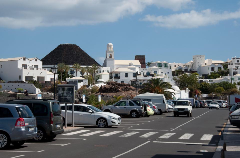 Street scene in Playa Blanca, Lanzarote, with white buildings, palm trees, and parked cars.