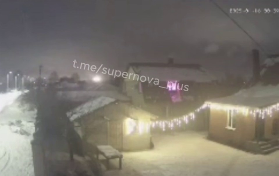 Nighttime security footage of a snow-covered area with buildings and lights.