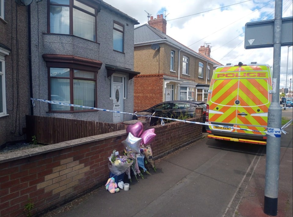 Crime scene photo showing a house with police tape and a police van; memorial flowers and stuffed animals are in the foreground.