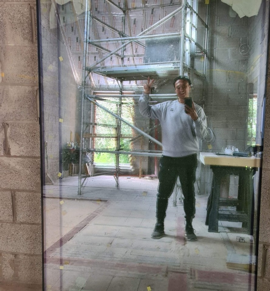 Reflection of a man in a grey sweatshirt taking a selfie in front of a large pane of glass in an unfinished room.