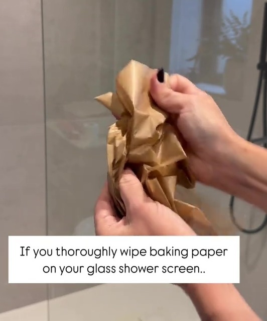 Hands wiping baking paper on a glass shower screen to prevent streaks.