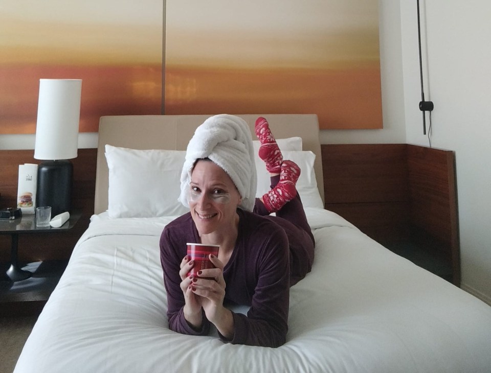 Woman in pajamas and eye mask relaxing on a hotel bed with a cup.