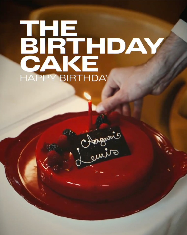 A red birthday cake with a single lit candle and the name "Lewis" written in icing.