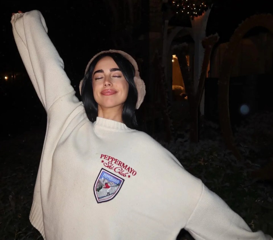 Woman in cream sweater with "Peppermayo Ski Club" logo, eyes closed, arm raised.