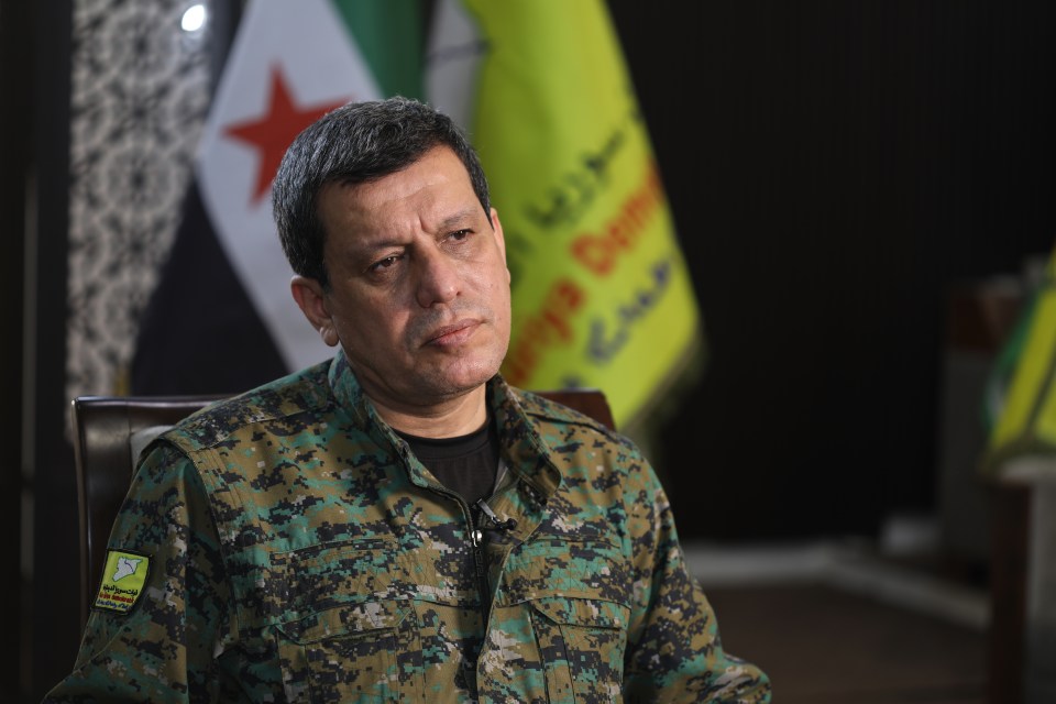 A man in camouflage uniform sits in front of Syrian flags.