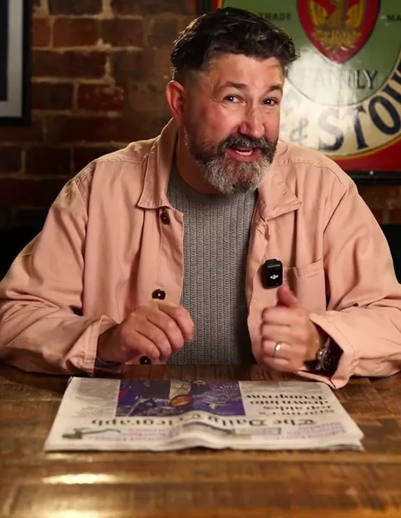 Man in pink shirt sitting at a table, looking at a newspaper.