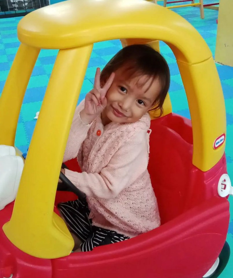 Young girl making peace sign while sitting in a toy car.