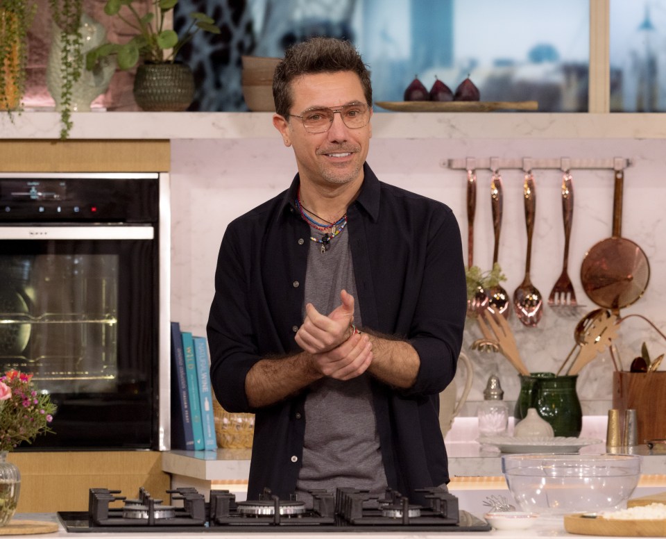 Gino D'Acampo, shirtless, holding a small Italian flag-colored knitted item, on a cooking show set.