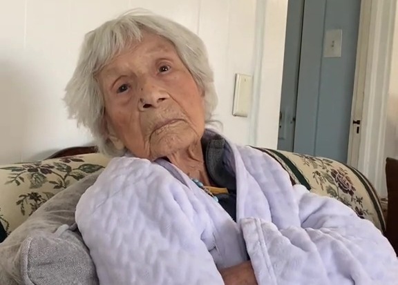 A 106-year-old woman in a lavender robe sits on a couch, arms crossed.  Text overlay asks what she used to wash her face and hair.