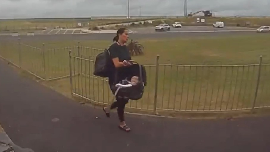 Woman carrying baby in car seat walks along path.