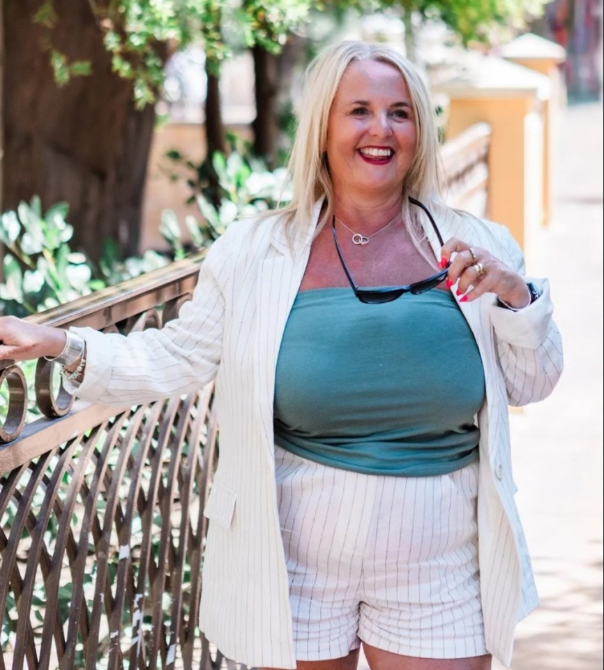 Woman in a white blazer and striped shorts smiles outdoors.