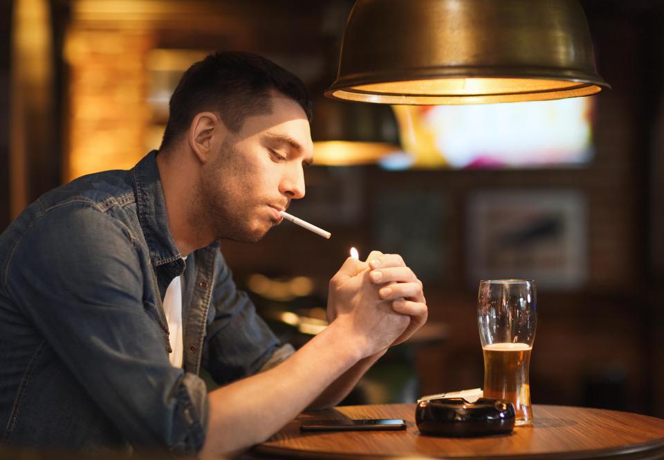 Man lighting a cigarette at a bar with a beer.