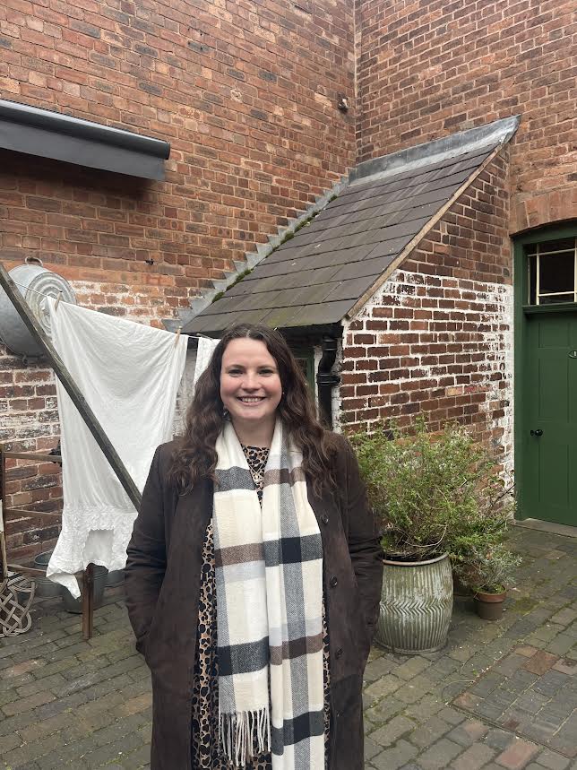 Woman standing in a brick courtyard wearing a brown coat and scarf.