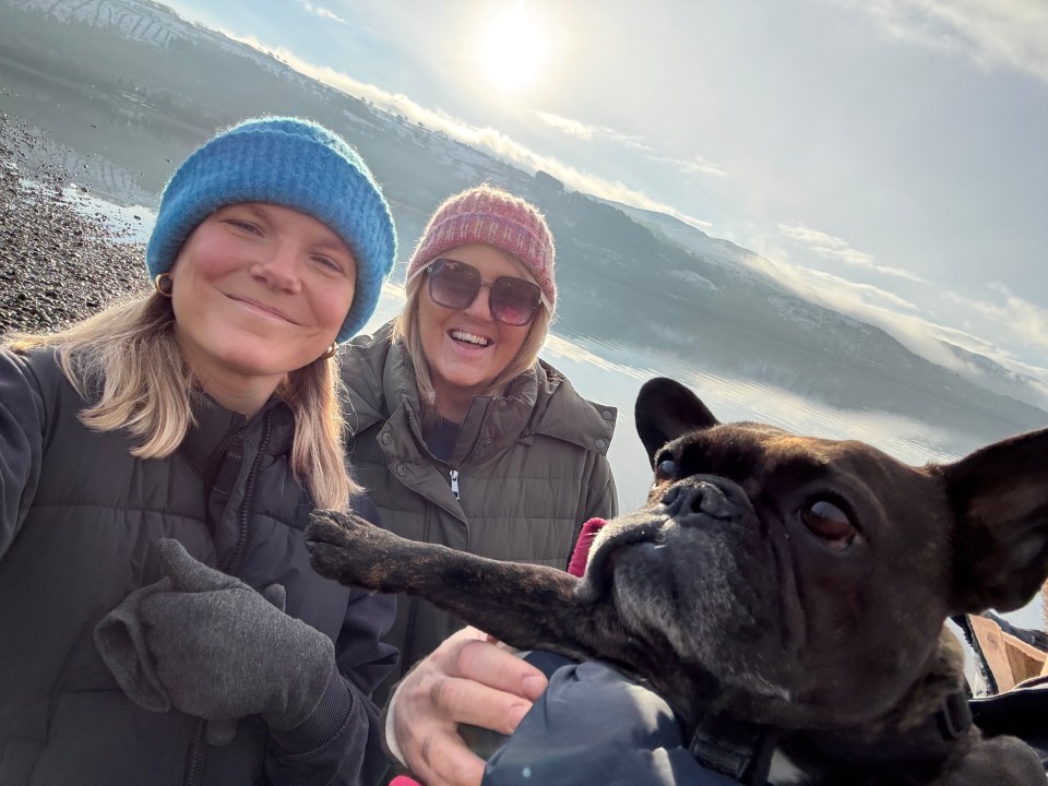 Two women and a French bulldog by a lake with snowy mountains in the background.