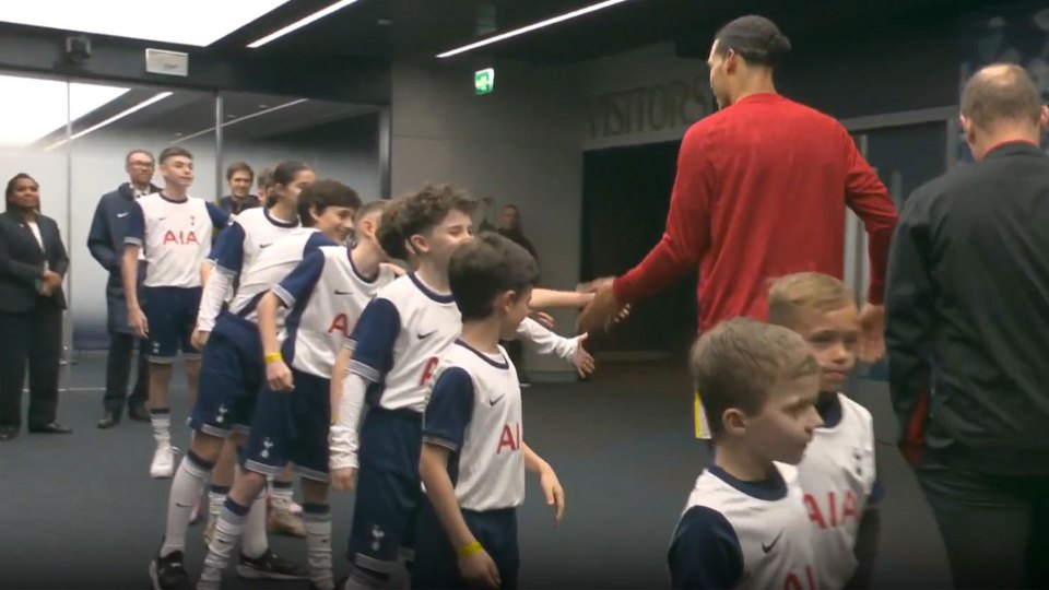 A tall man in a red shirt greets a line of children in soccer uniforms.