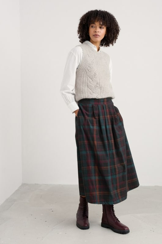 Woman wearing a light gray knitted vest, a pleated plaid midi skirt, and burgundy boots.