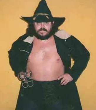 Portrait of a shirtless wrestler in a long black coat and large hat, holding metal restraints.