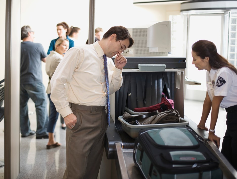 A businessman watches as a security guard inspects his luggage at an airport security checkpoint.