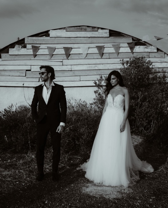 Black and white photo of a bride and groom posing in front of a rustic wooden structure.