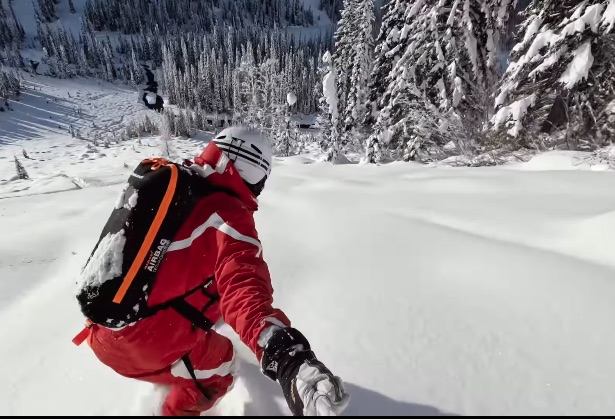 Snowboarder wearing a red jacket and airbag backpack riding down a snowy mountain slope.