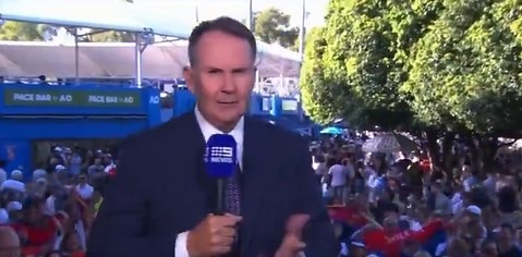 News reporter holding a microphone in front of a crowd at a tennis tournament.