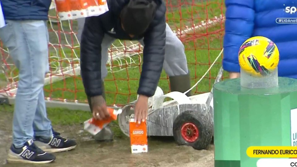 Person pouring liquid onto a small model car near a soccer goal and soccer ball.