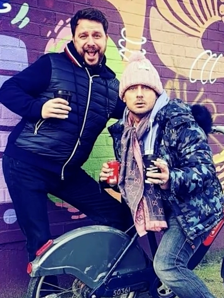 Two men standing by a graffitied wall, one holding a coffee cup, the other with two coffee cups and a bicycle.